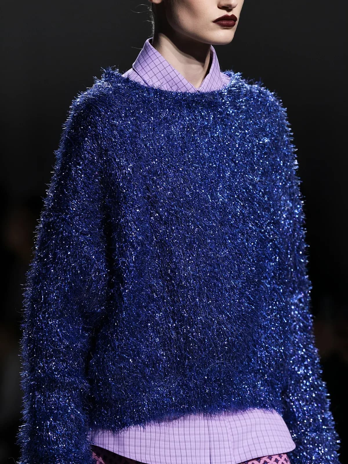 A model in a shimmering blue tinsel sweater over a lilac checked shirt, against a dark background.